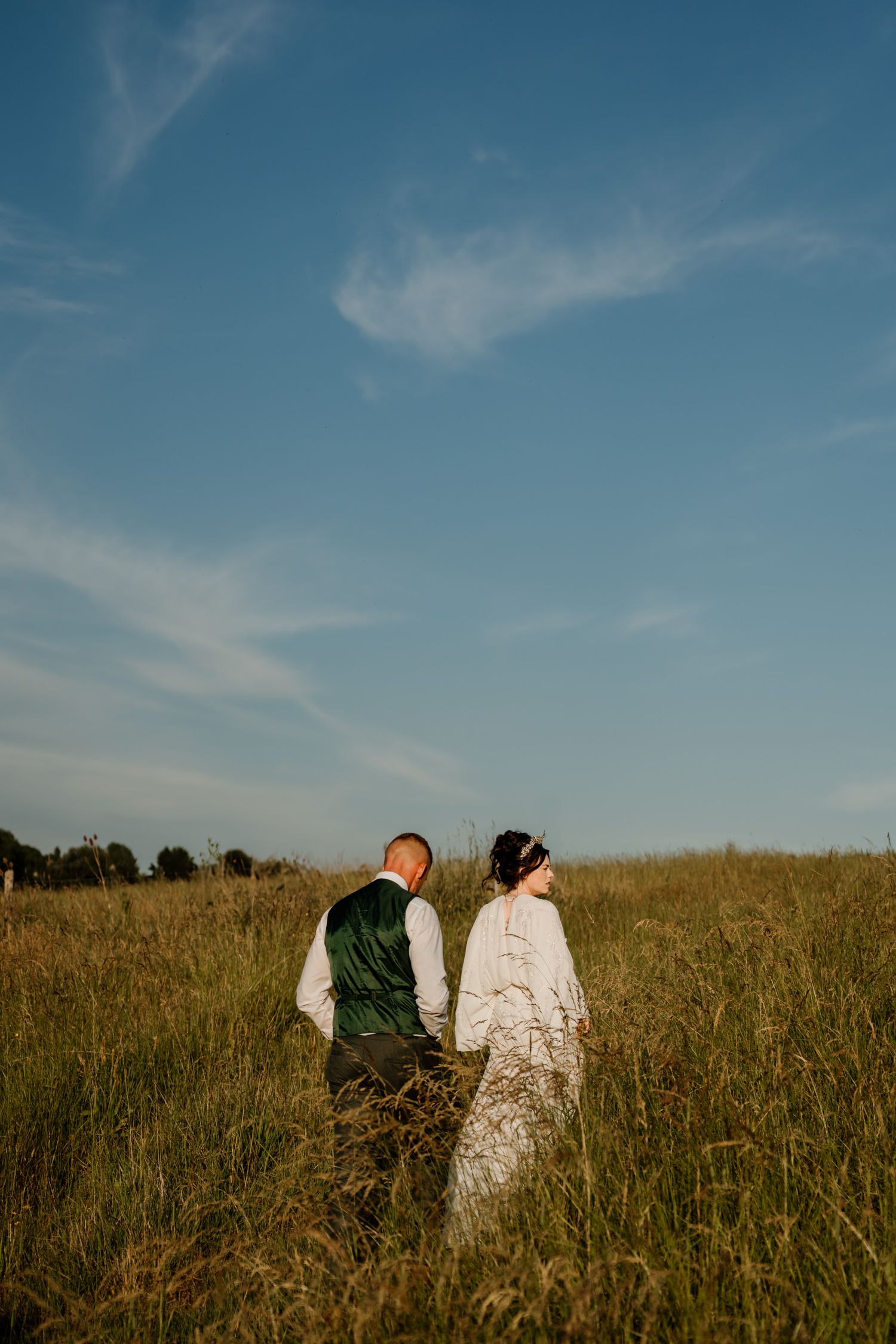 Newlyweds walk through tall grass under bright blue skies during their countryside wedding photoshoot.