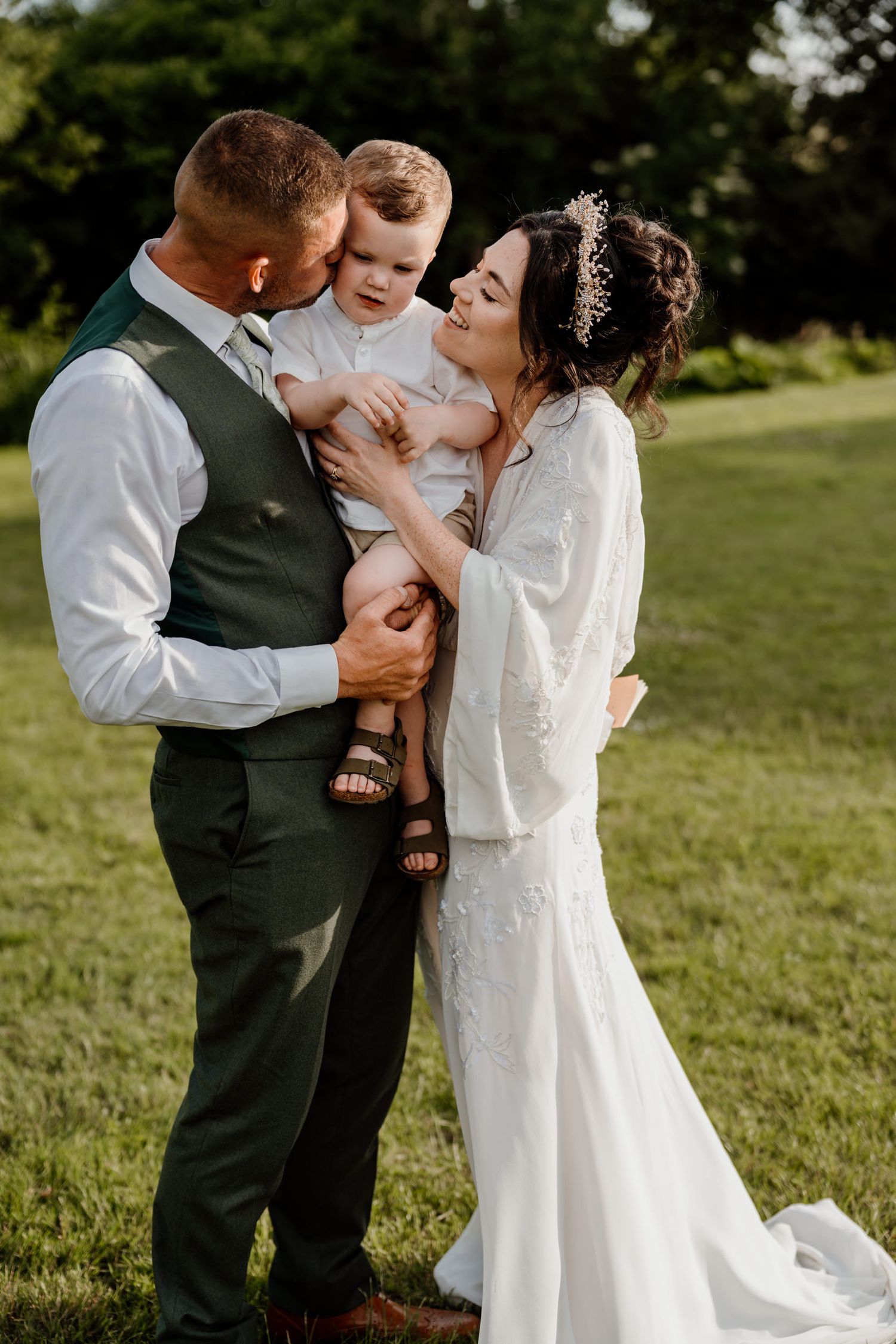 A tender family moment captured outdoors during a wedding celebration in a green field.