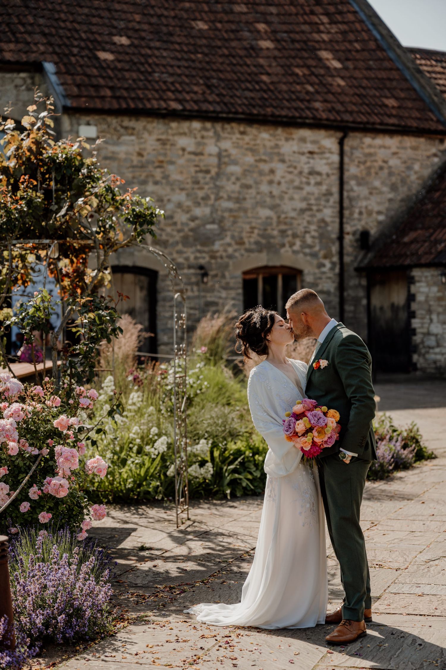 Couple poses in a rustic courtyard garden with blooming flowers and stone walls on a sunny day.