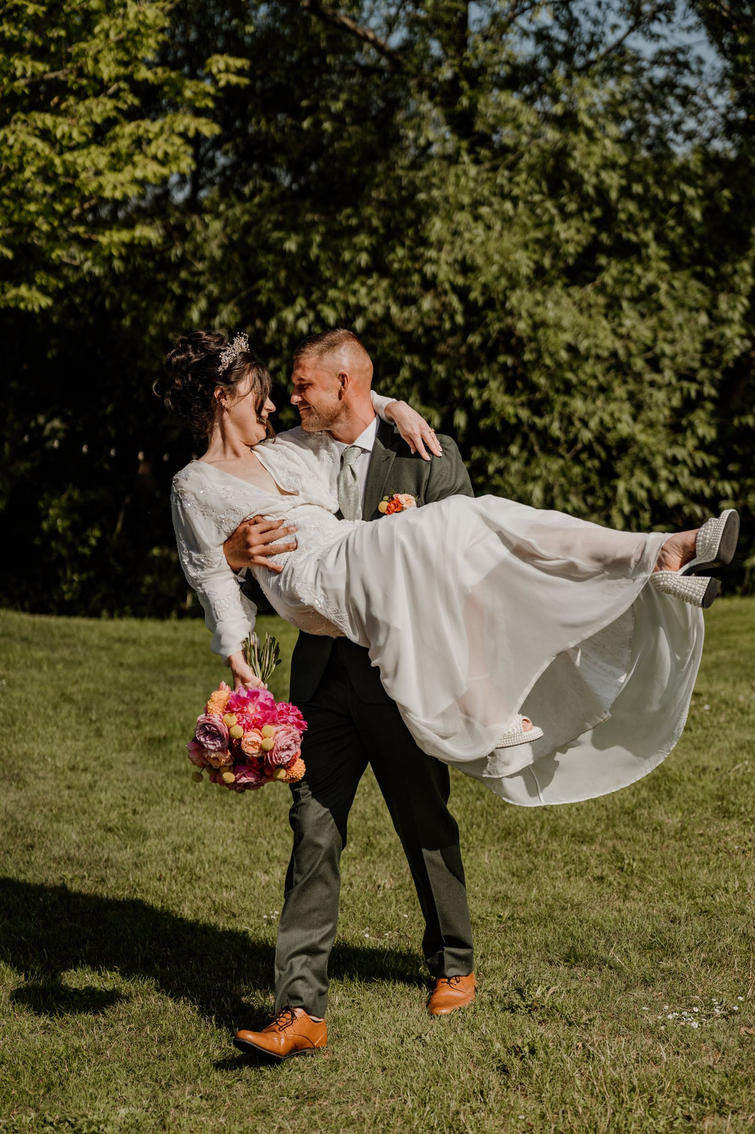 A playful wedding couple poses in a sunlit garden with the groom lifting the bride's flowing white dress.