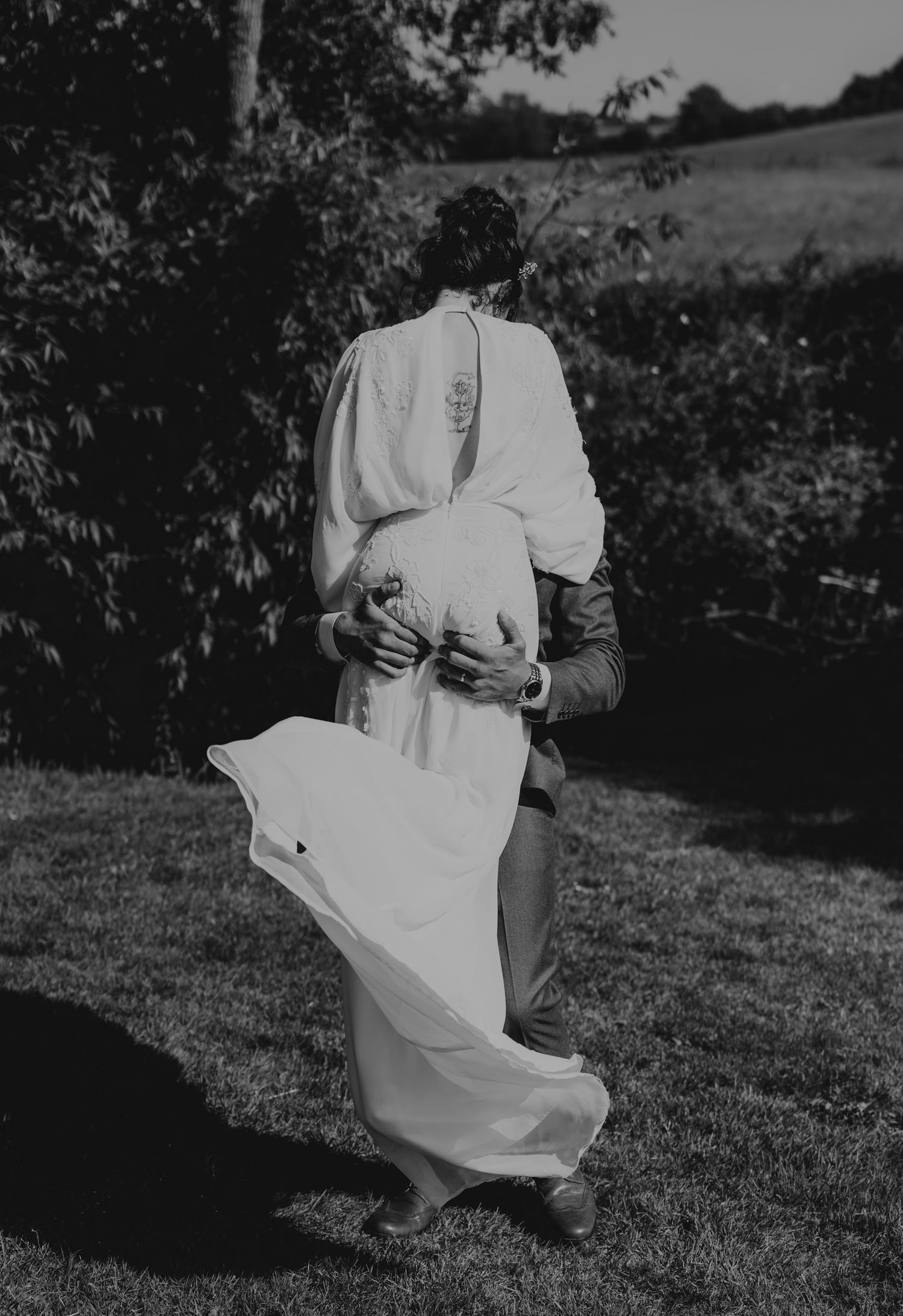 Black and white photo of newlyweds dancing playfully outdoors with the bride's dress flowing in motion.