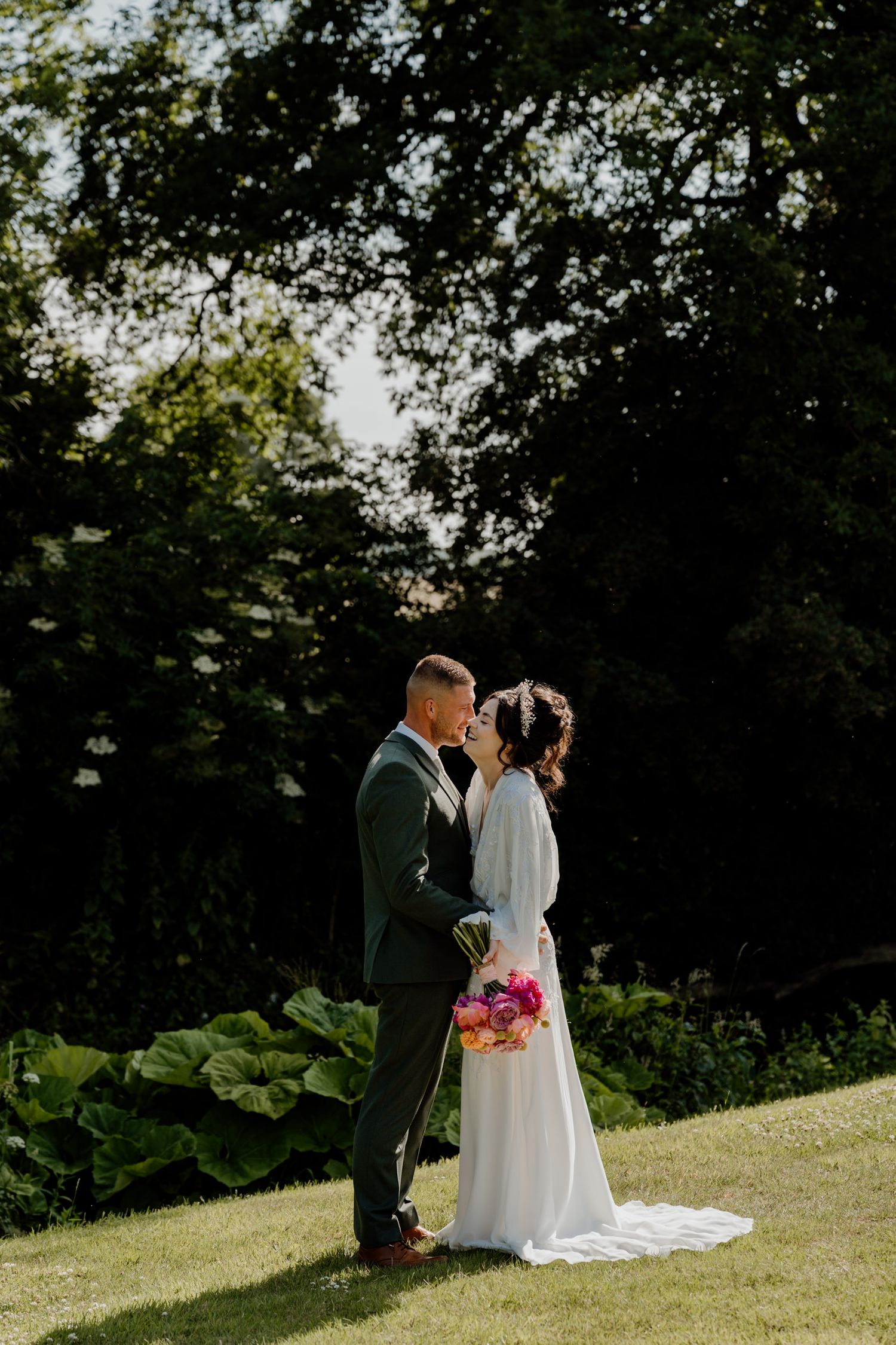 Newlyweds embrace on a grassy slope with trees in the background while the bride holds a bright pink bouquet.