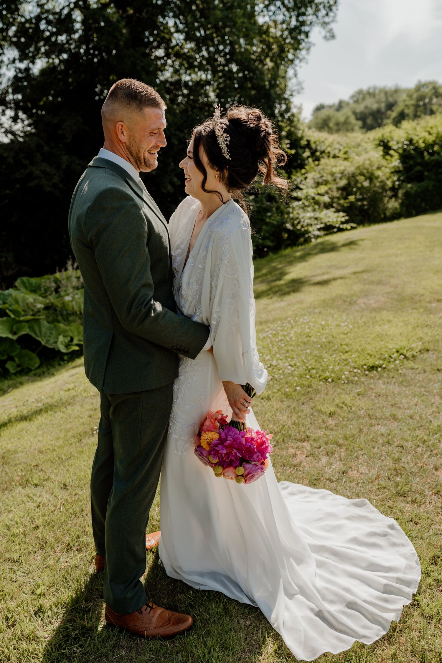 A couple in wedding attire share an intimate moment on a sunlit grassy hillside with flowing white dress and vibrant bouquet.