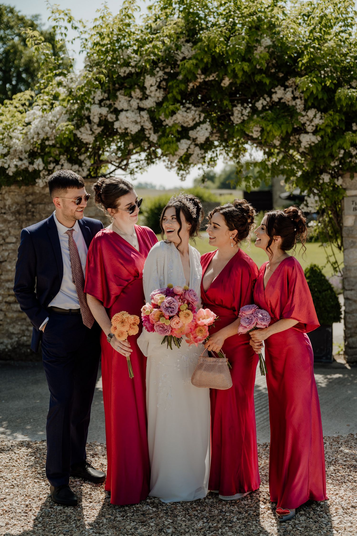 Wedding party in red bridesmaid dresses and white bridal gown pose together outdoors under trees.