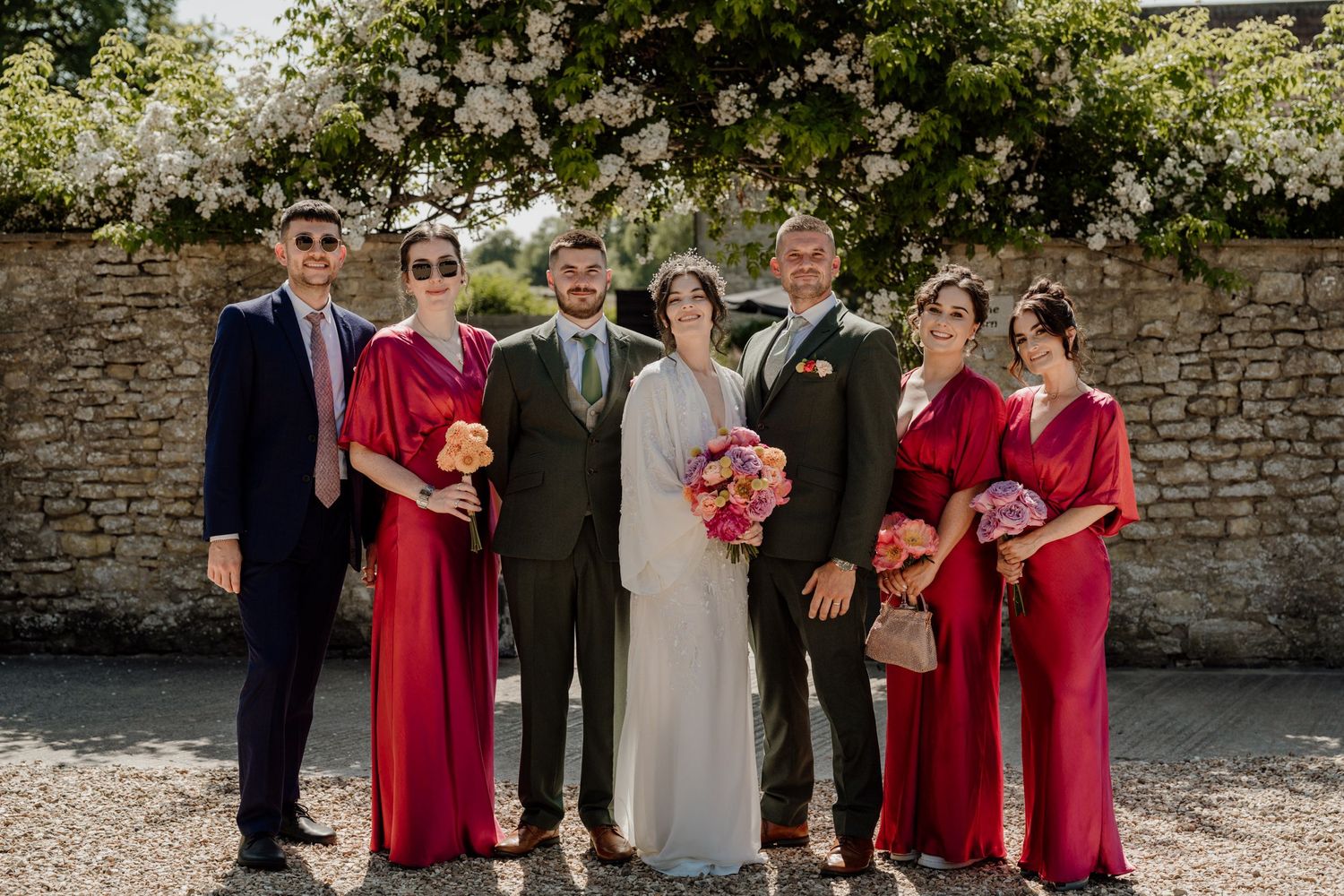 Wedding party poses outdoors against rustic stone wall wearing red bridesmaid dresses and formal attire.