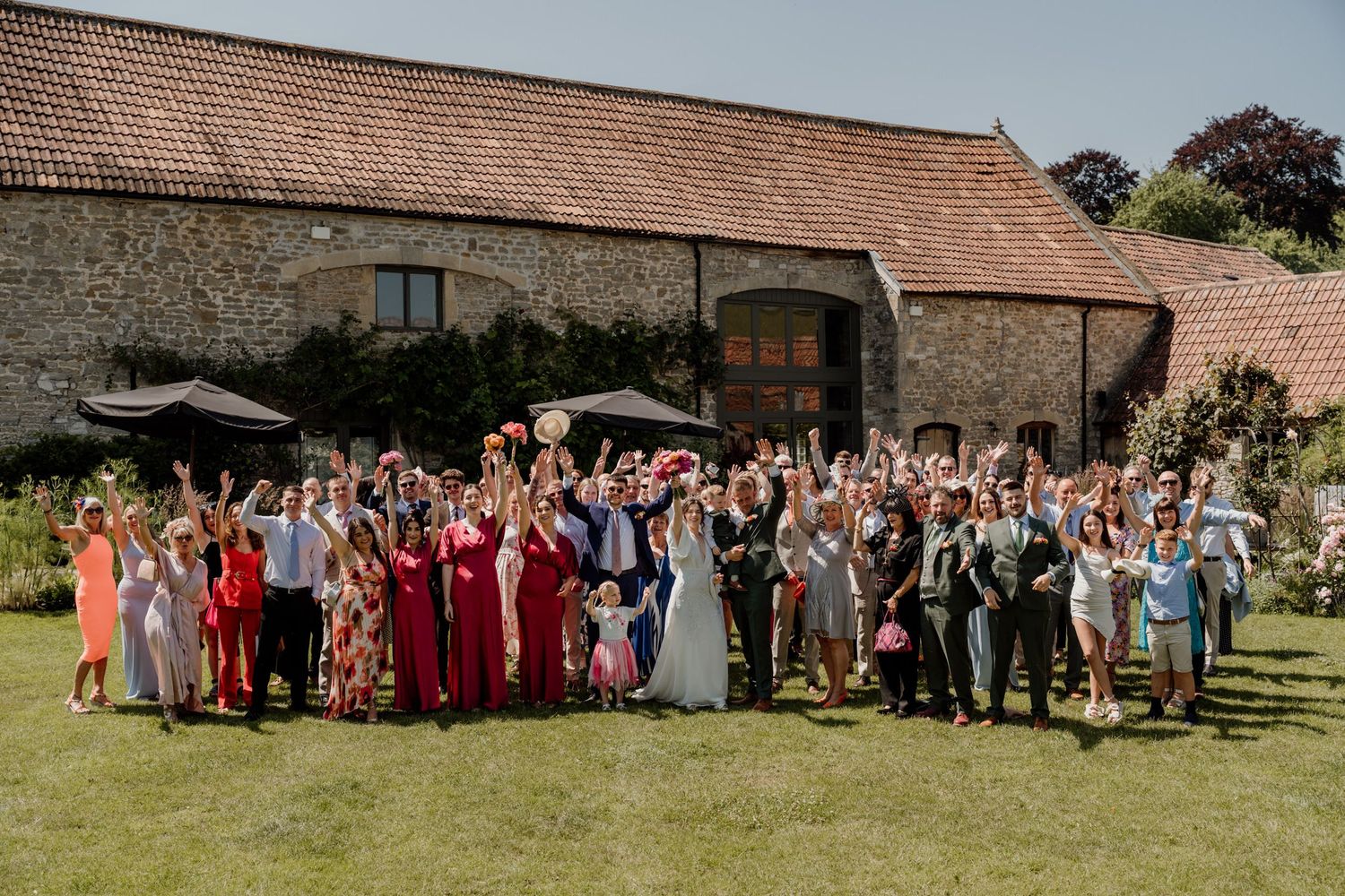 Large wedding party poses for a group photo outside a rustic stone barn on a sunny day.