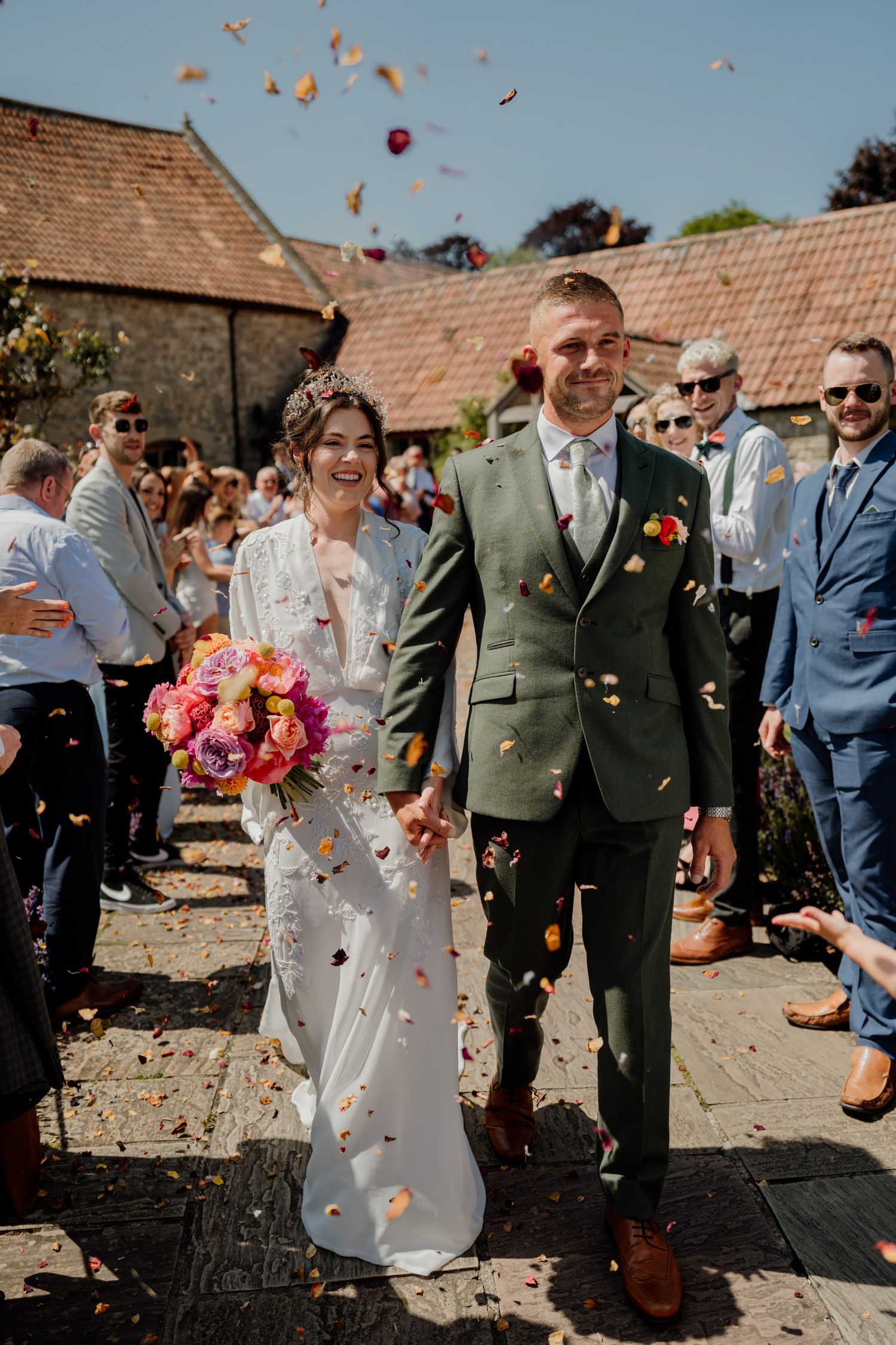 Wedding guests throw colorful autumn leaves and rose petals while celebrating on a sunny day at a rustic outdoor venue.