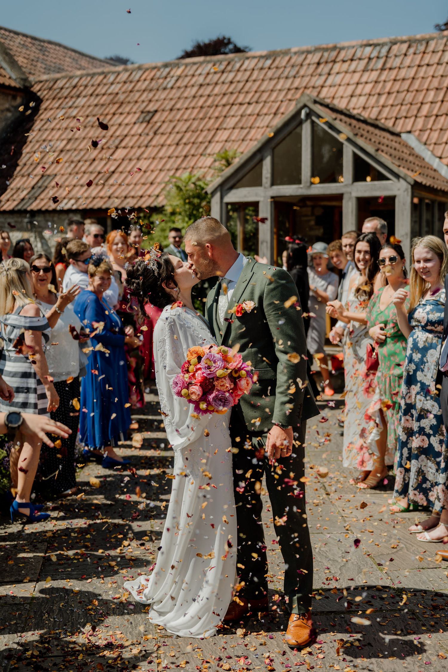 Wedding guests shower the couple with colourful confetti outside  at Priston Mill in Bath.