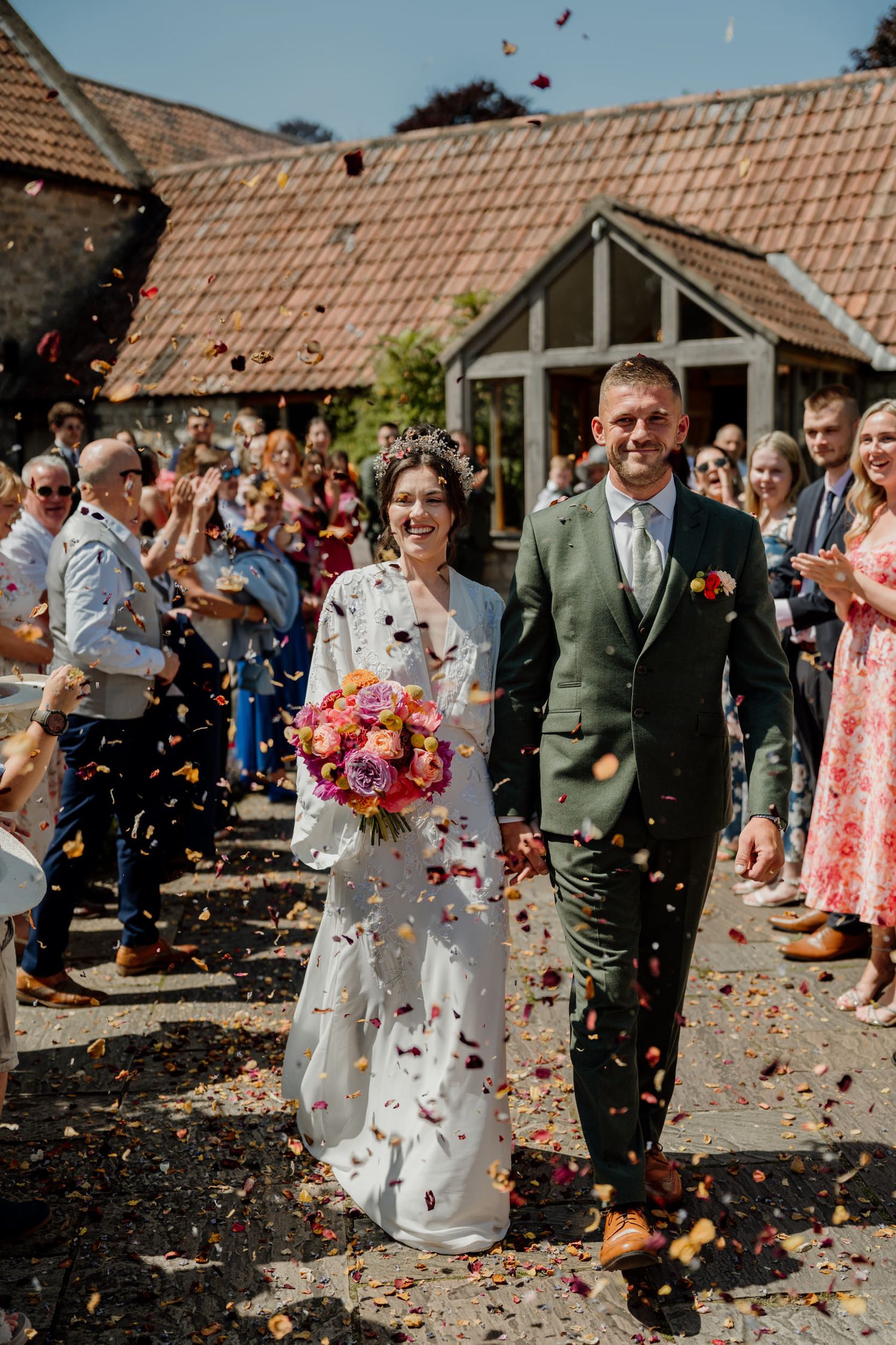 Wedding guests shower the newlyweds with colorful flower petals and confetti outside a rustic venue.
