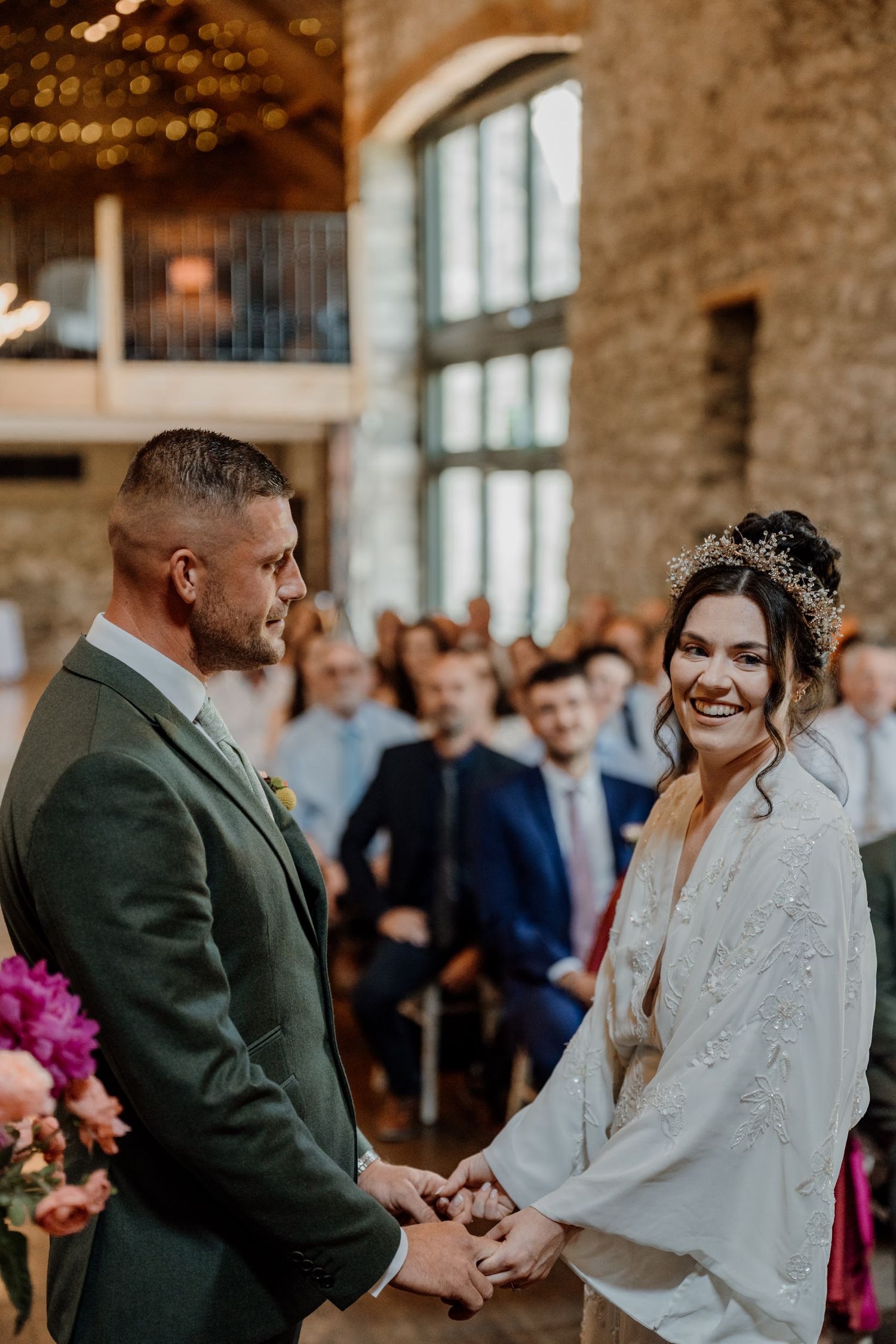 A joyous wedding ceremony takes place inside a stone-walled barn venue with large windows.
