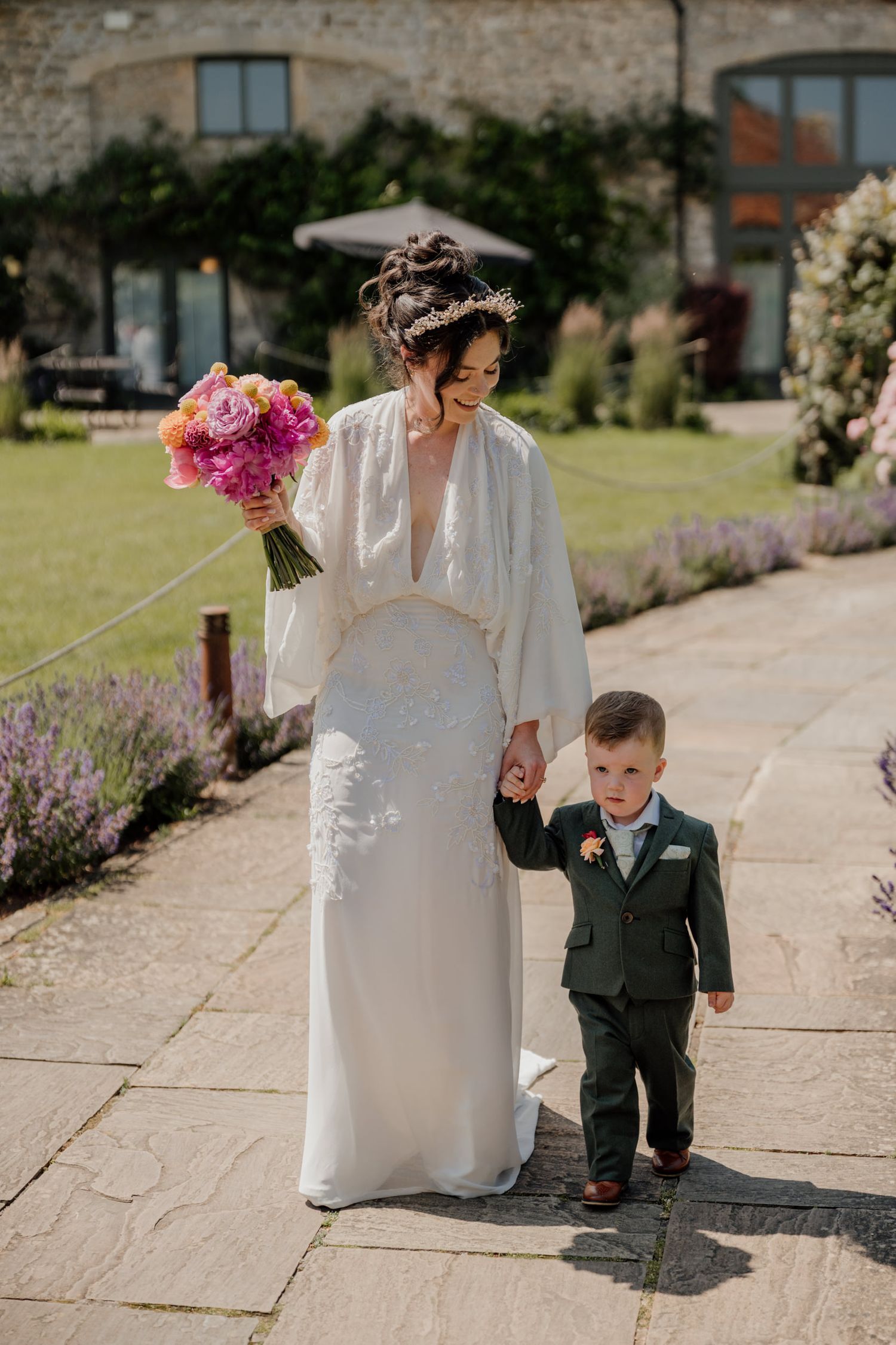 Small child walking alongside person in white dress on stone pathway surrounded by flowers.
