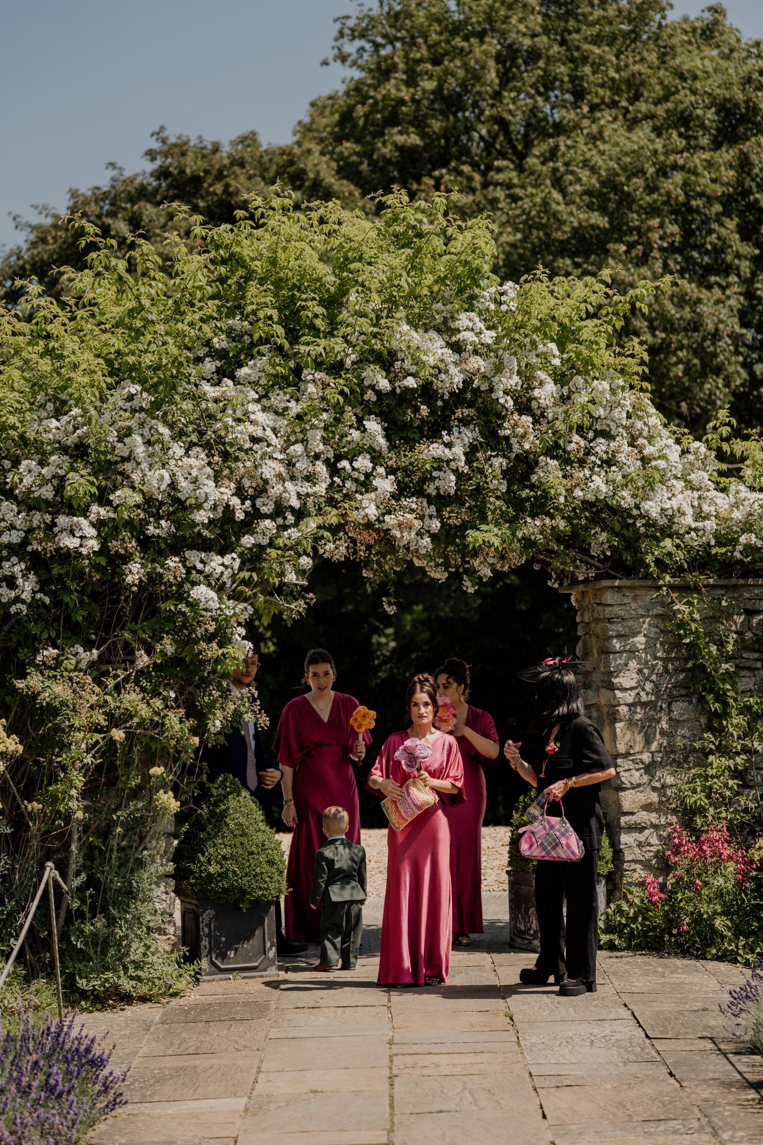 Wedding party in burgundy dresses gathered in a beautiful garden setting with white climbing roses at Priston Mill in Bath.