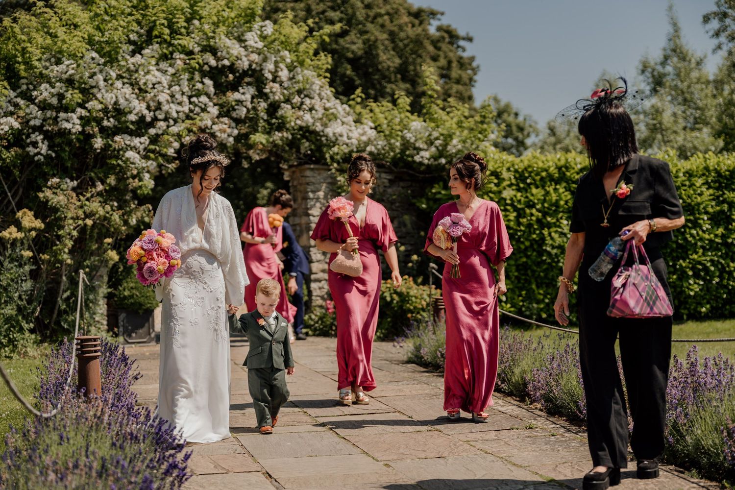 Wedding party members in pink attire walking through a garden pathway.