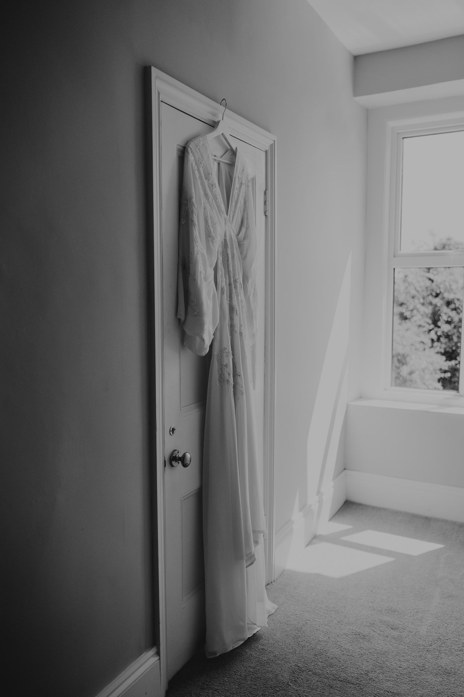 White wedding dress hanging on door in bright sunlit room with minimal decor.