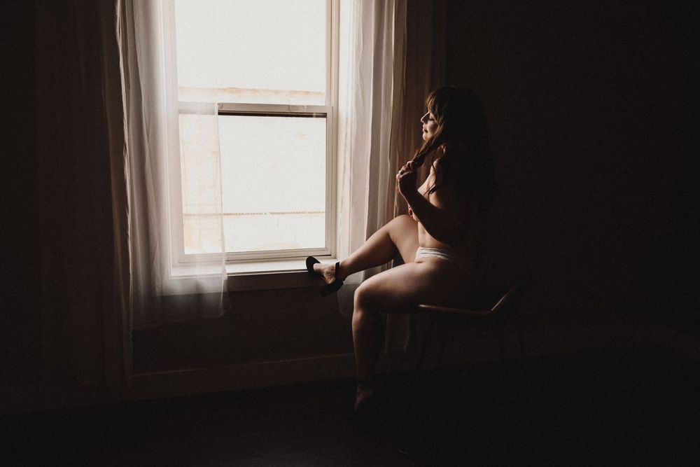 A woman sits by a sunlit window in soft natural light, captured in a moody boudoir photography session.