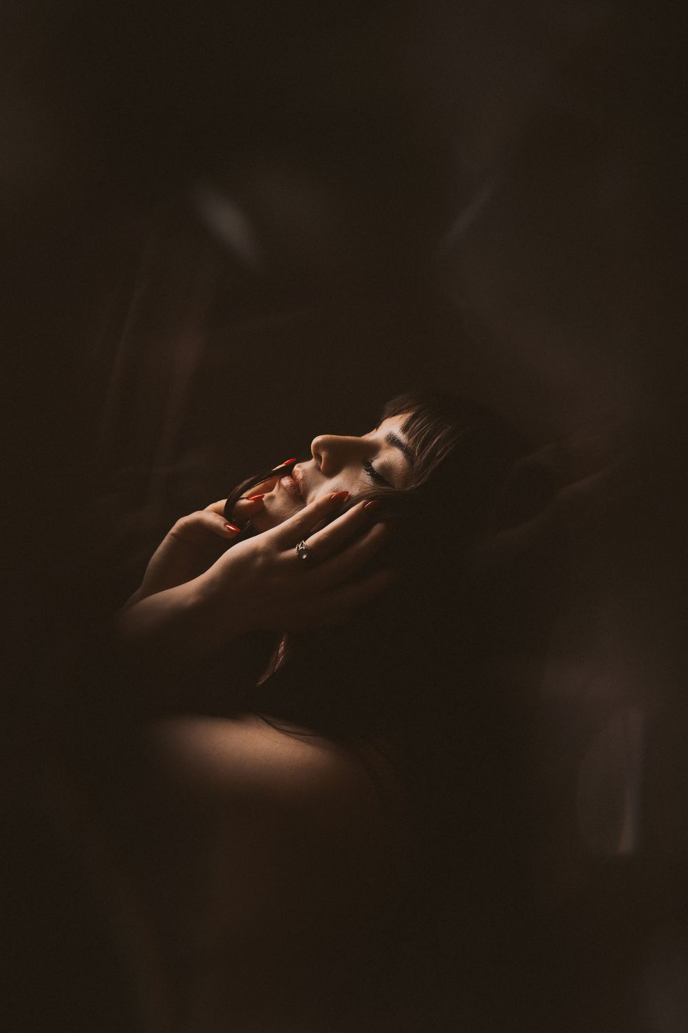 A woman with closed eyes tilts her head back in dramatic low-key lighting, hands gently cradling her face.