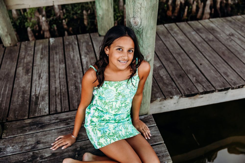 Young person in a mint green floral dress sits on wooden deck with rustic backdrop.