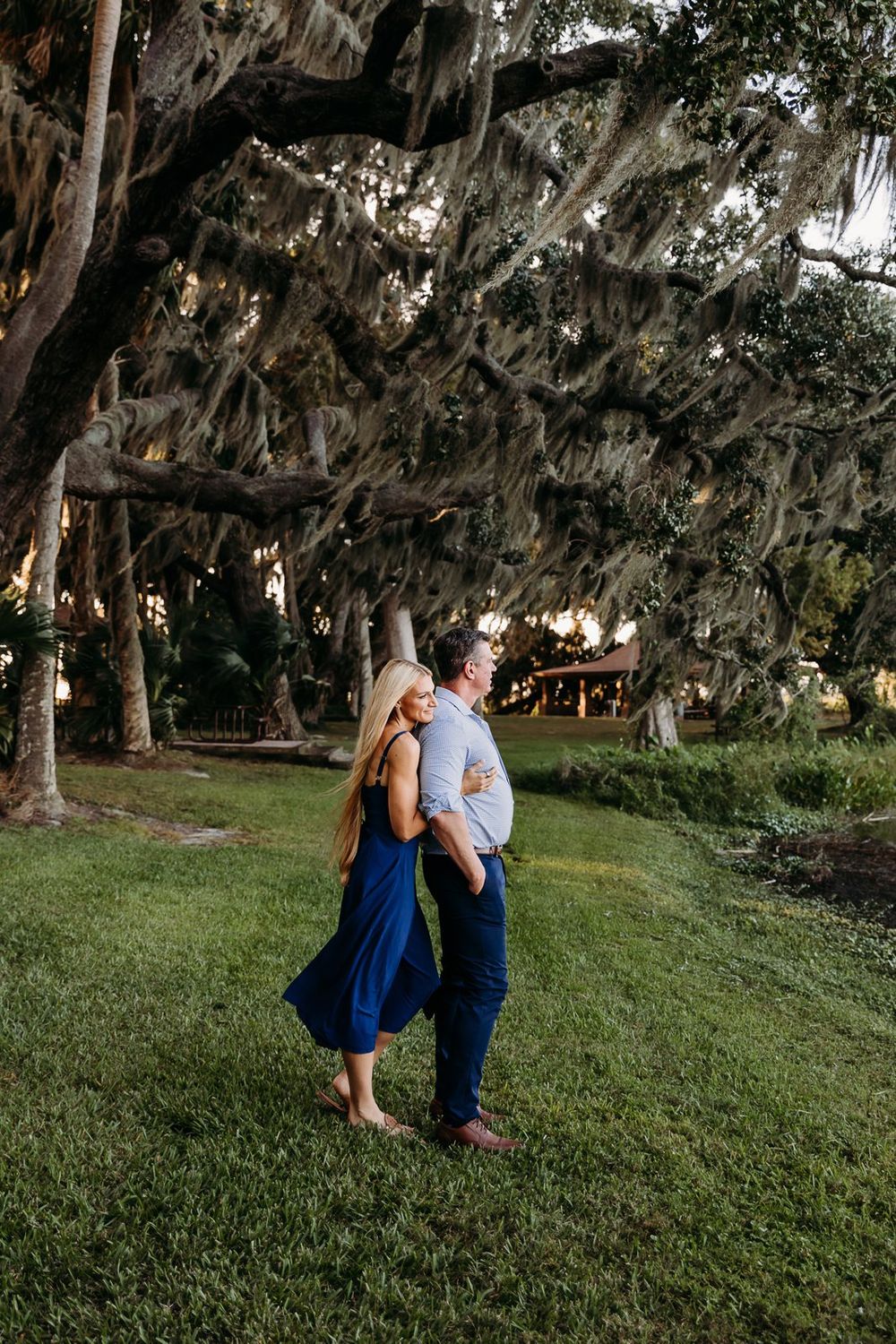 A romantic couple shares tender moments under Spanish moss-draped trees in a lush green park setting at sunset.