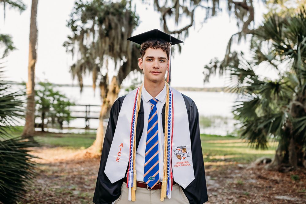 A graduate in academic regalia with honors stoles and graduation cap poses outdoors among palm trees and Spanish moss.