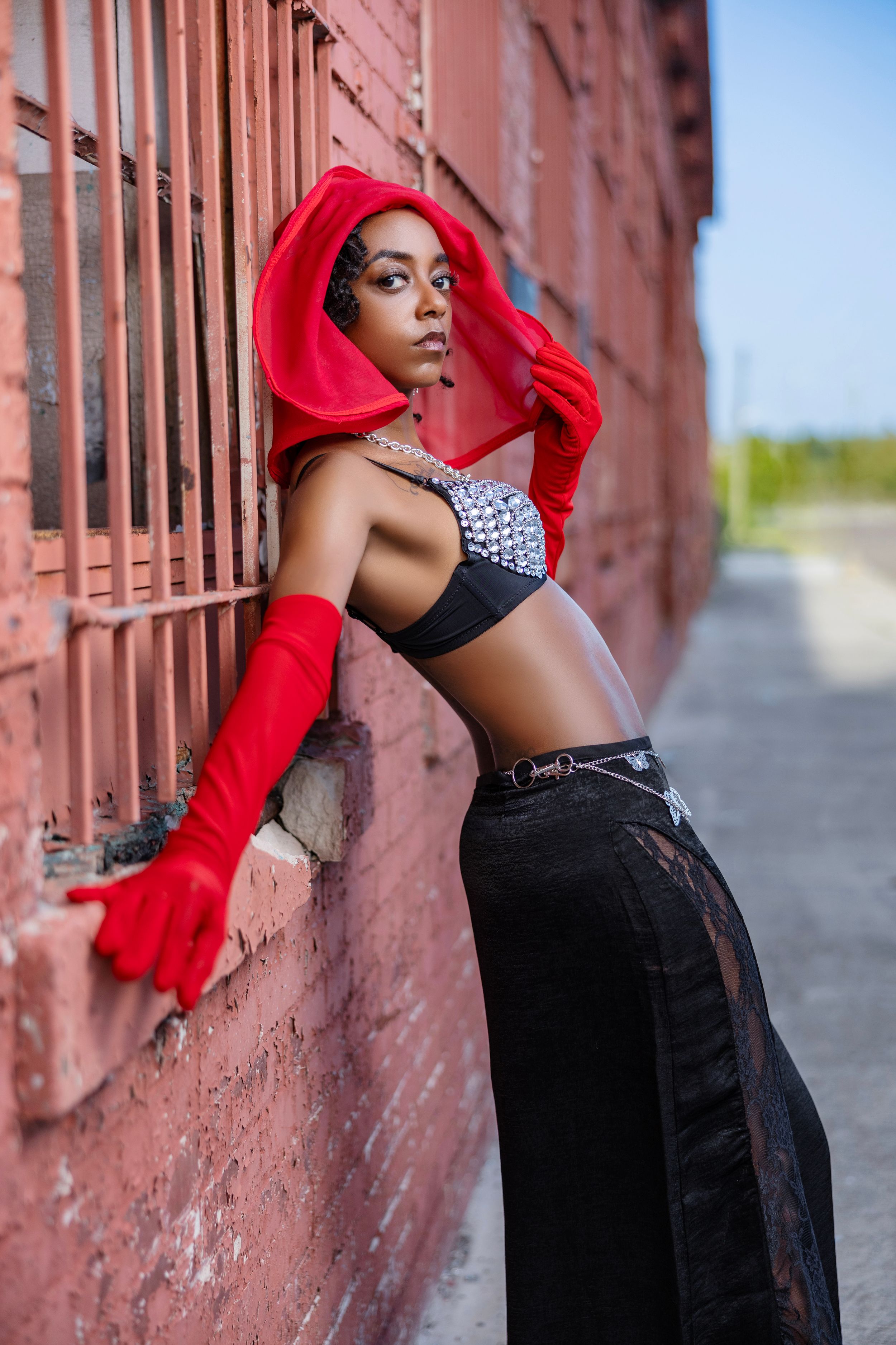 Fashion model in red hood and black outfit poses against pink wall in dramatic urban photoshoot.
