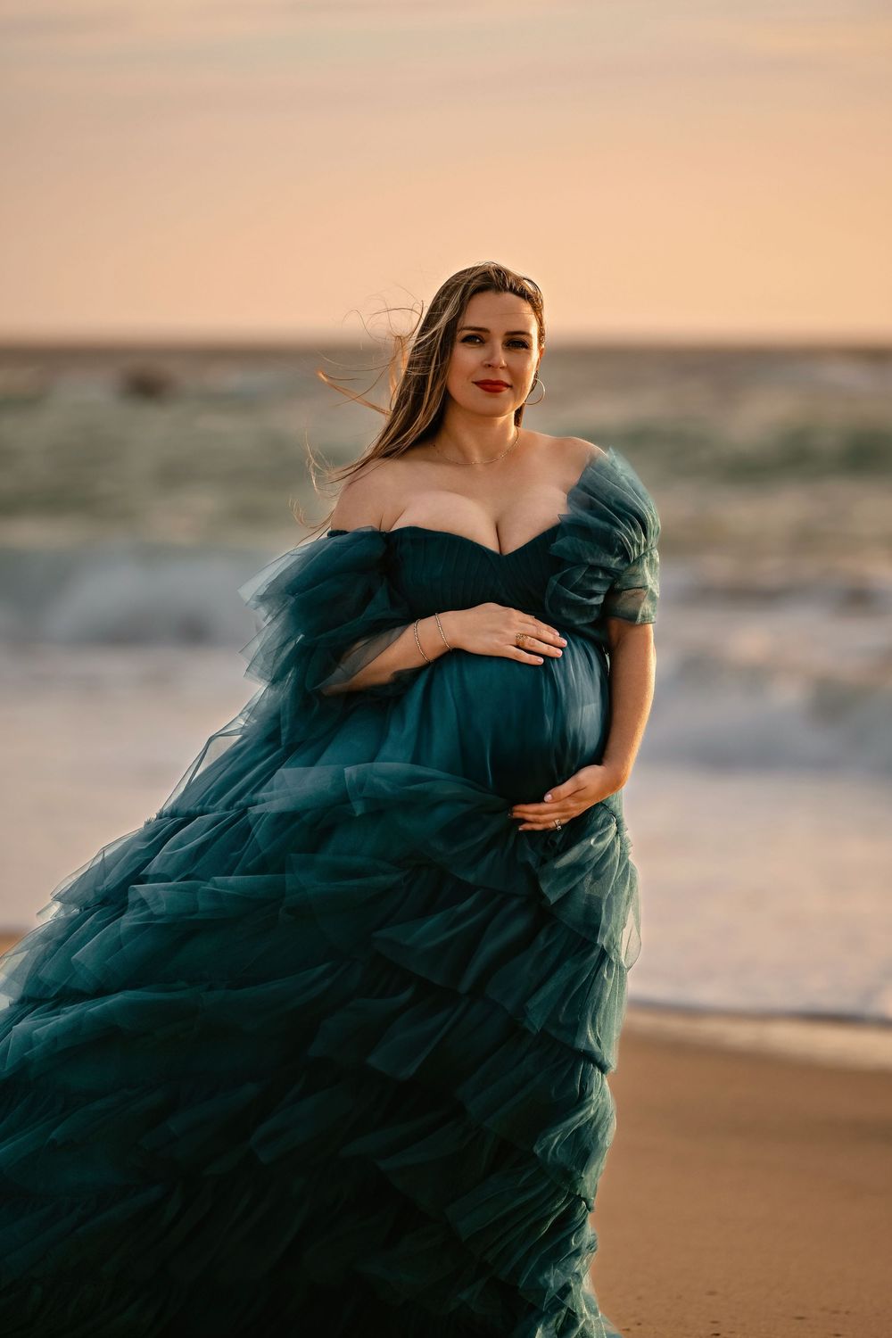 Flowing teal gown maternity photos on beach at sunset with dramatic fabric movement.