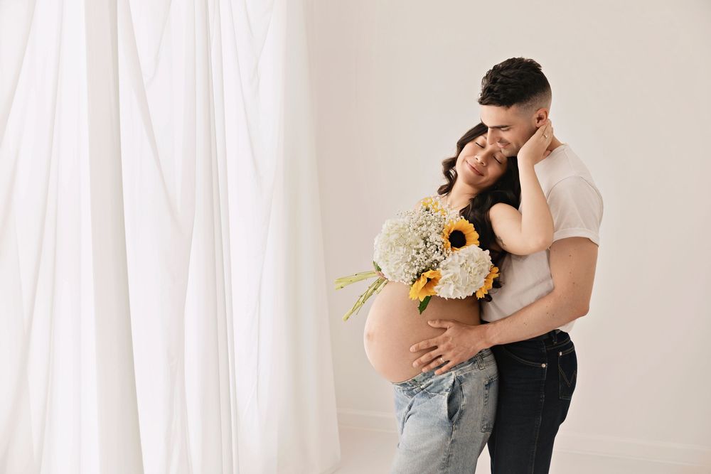 Pregnant woman in jeans holding sunflowers embraced by partner near white curtains in a maternity photo shoot.