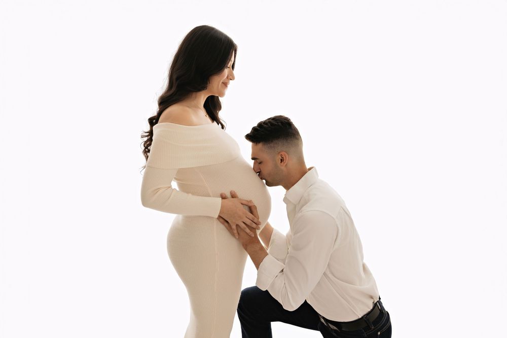 Man kneeling and kissing pregnant woman's belly during a maternity photo shoot on white background.