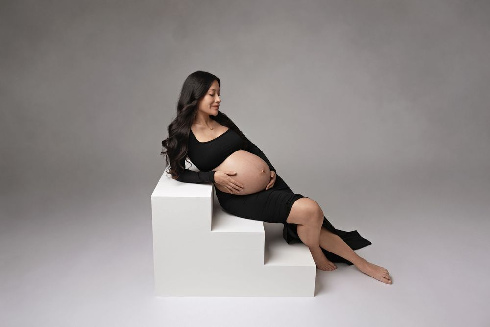 Pregnant woman in black dress sitting on white steps, cradling baby bump in professional maternity photo studio.
