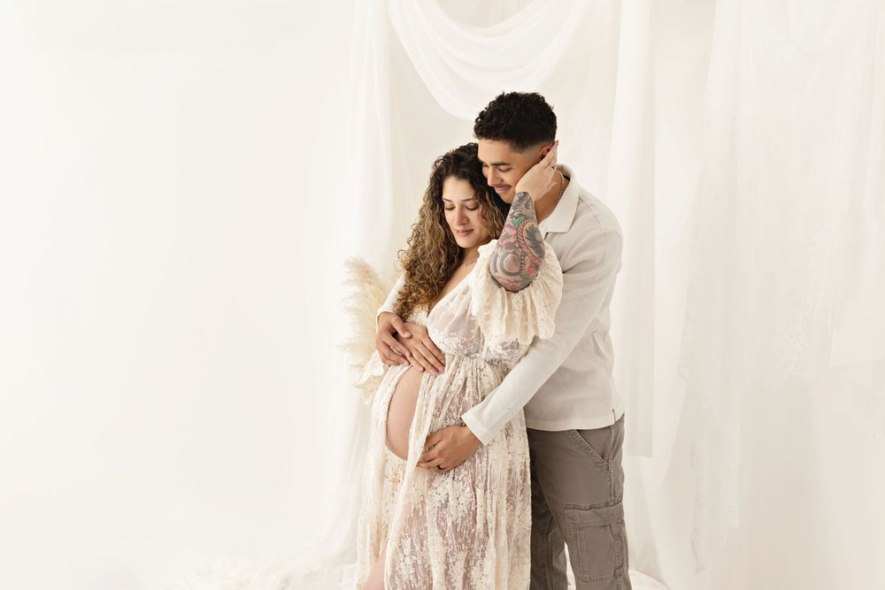 Couple in a maternity photo shoot, woman in lace dress cradling baby bump, man embracing her in white studio.