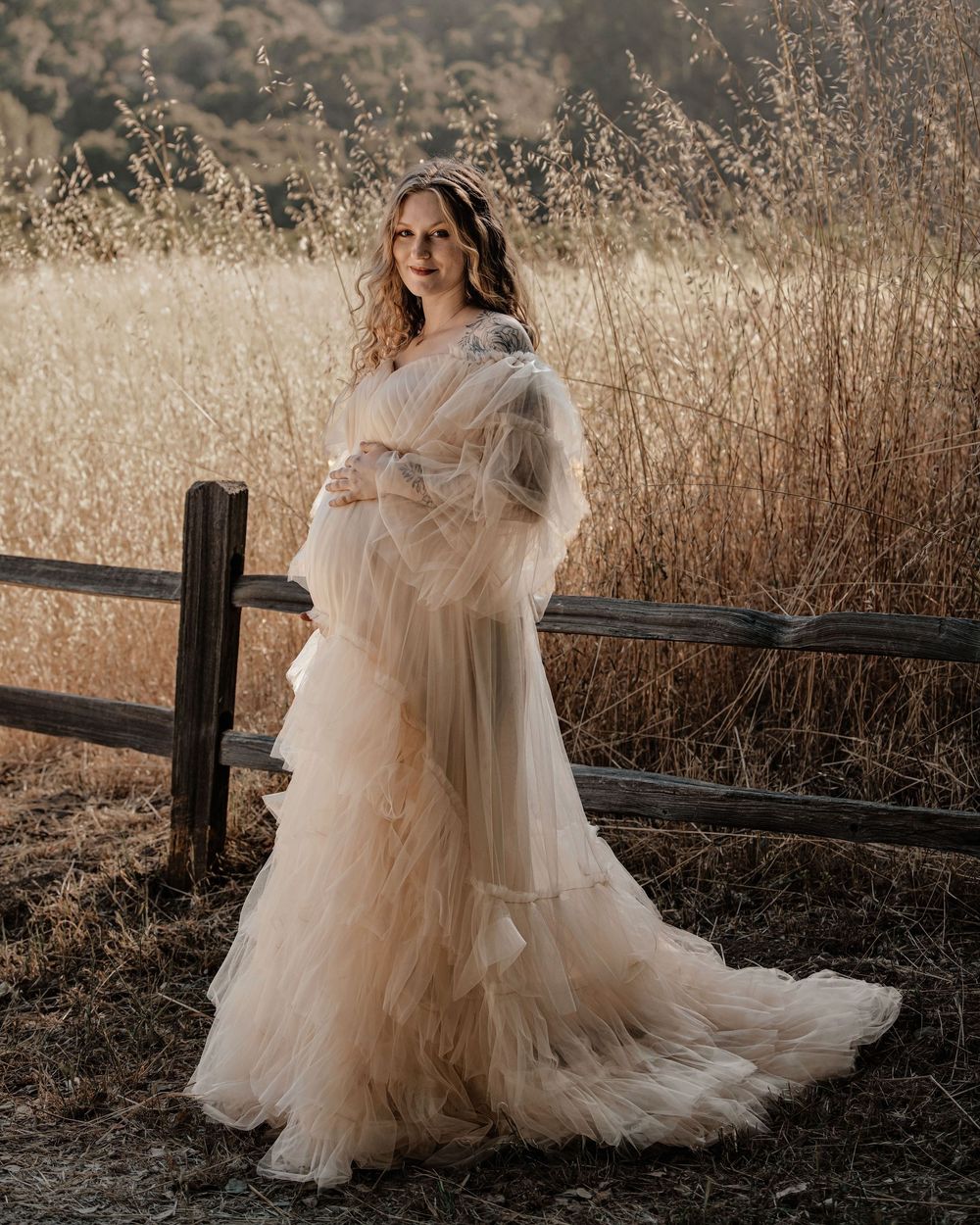 Ethereal maternity photos in flowing tulle dress against rustic wooden fence at golden hour.