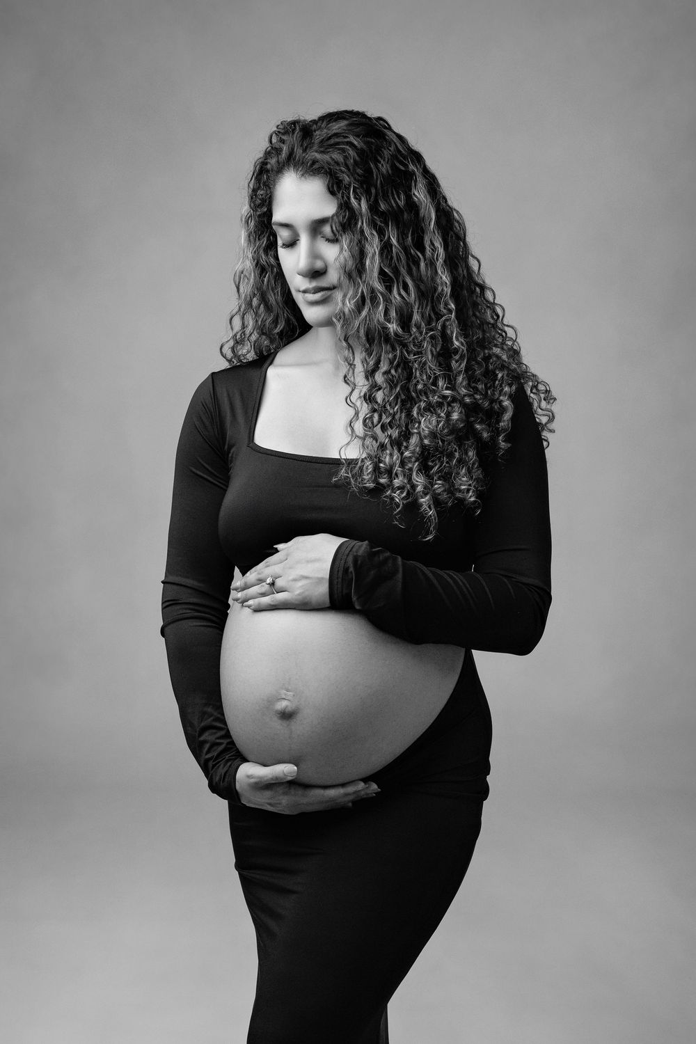 Black and white portrait of a pregnant woman with curly hair cradling her baby bump in a black dress.
