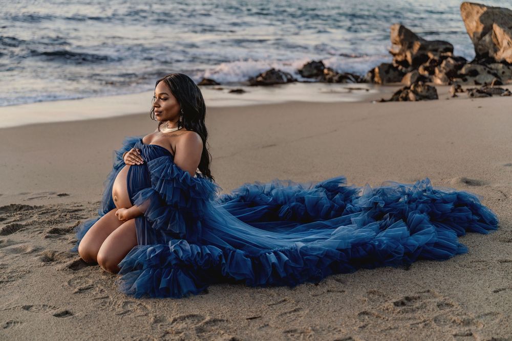 Dramatic coastline photoshoot features pregnant woman in a flowing royal blue dress against ocean waves and sandy beach at golden hour.