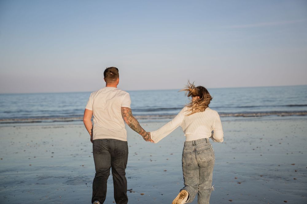 Couple holding hands walking on a sandy beach toward the calm ocean under a clear blue sky.