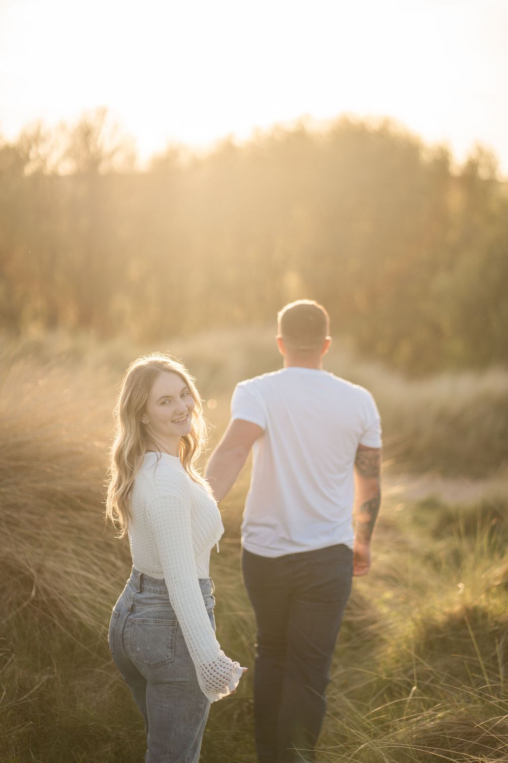 Couple holding hands in golden sunlit field, woman in white sweater and jeans smiling over shoulder.