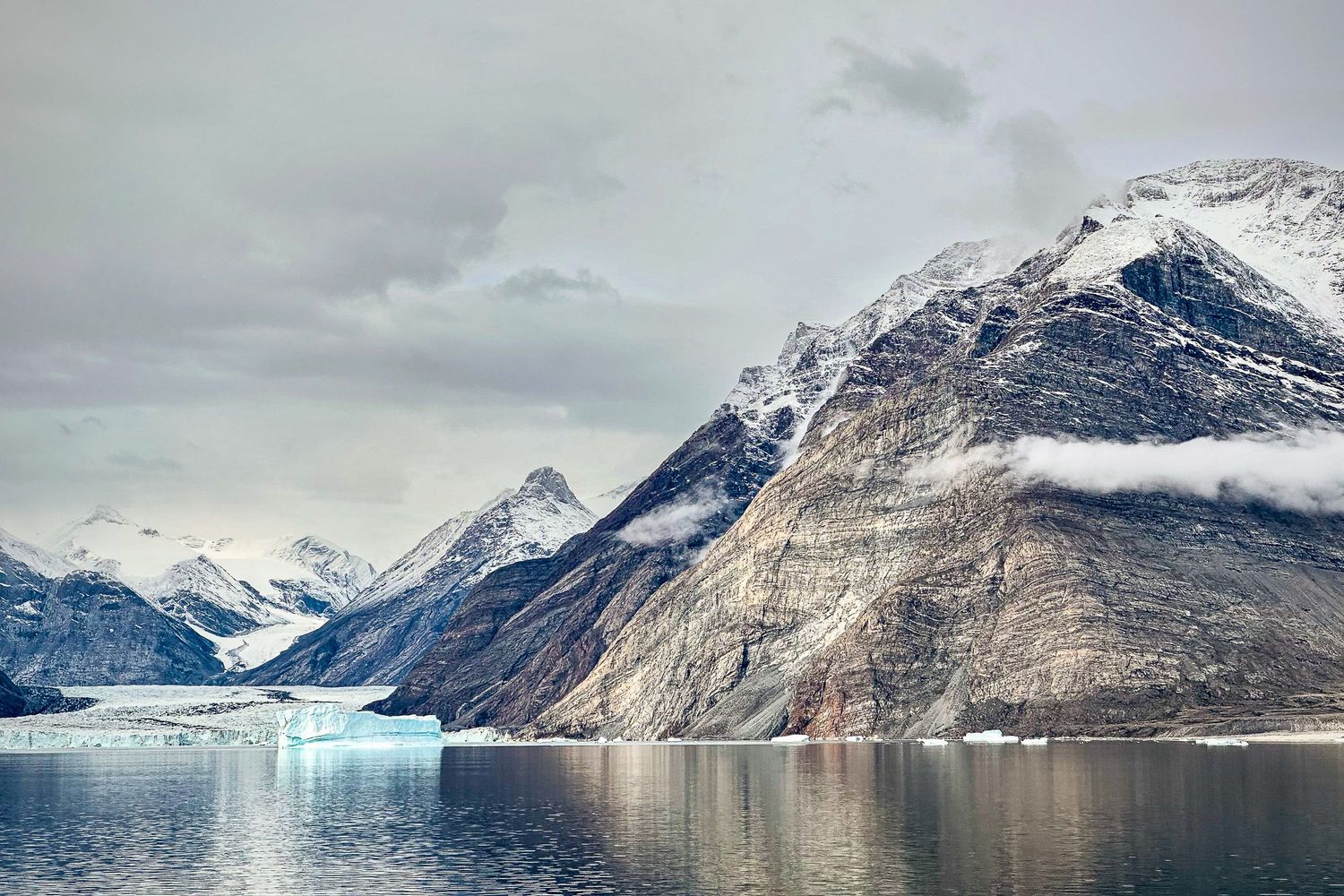 Snow-capped mountain peaks tower above a serene glacial fjord with icy waters reflecting the dramatic landscape.