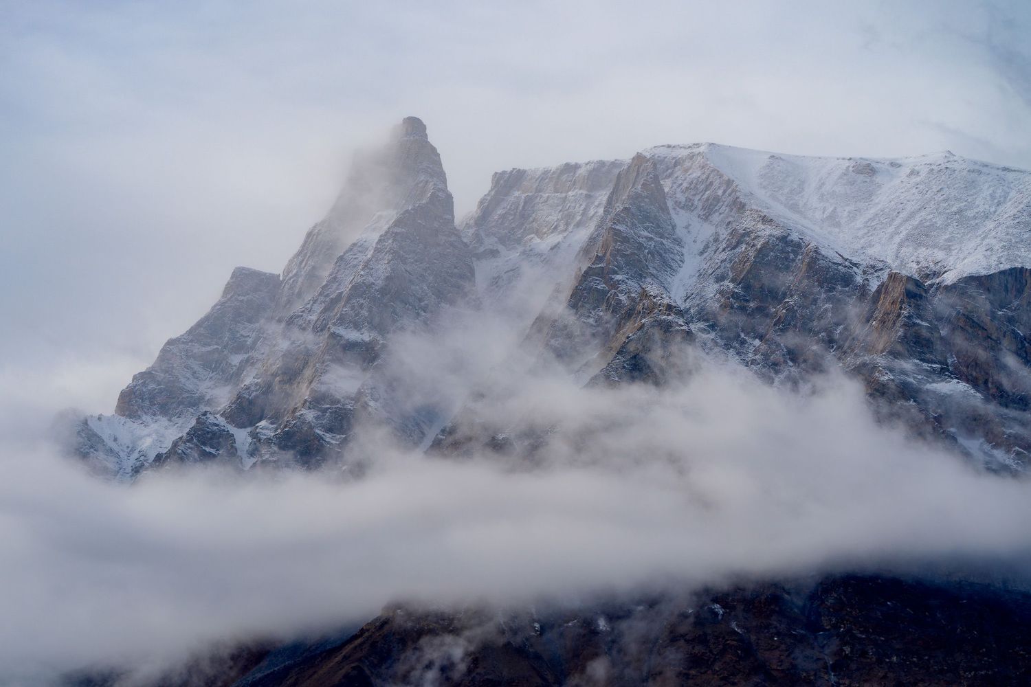 Snow-capped mountain peaks shrouded in misty clouds against a moody blue sky near a mountain range.