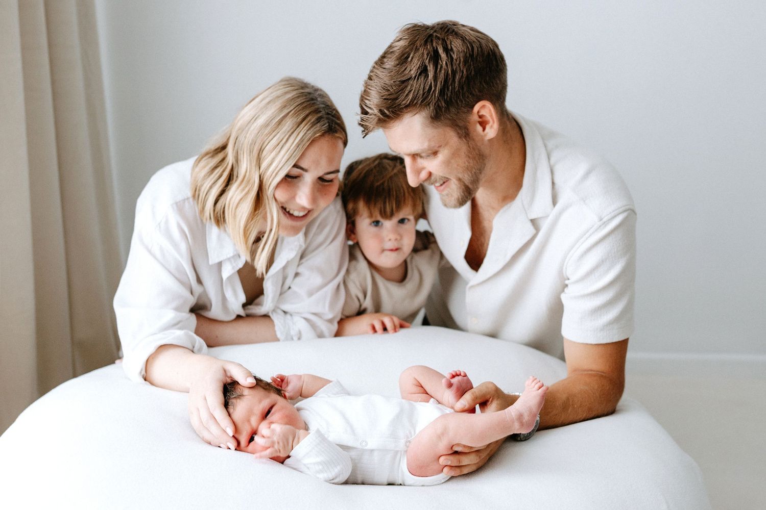 A family lies together on a white bed sharing tender moments with their newborn baby at home.