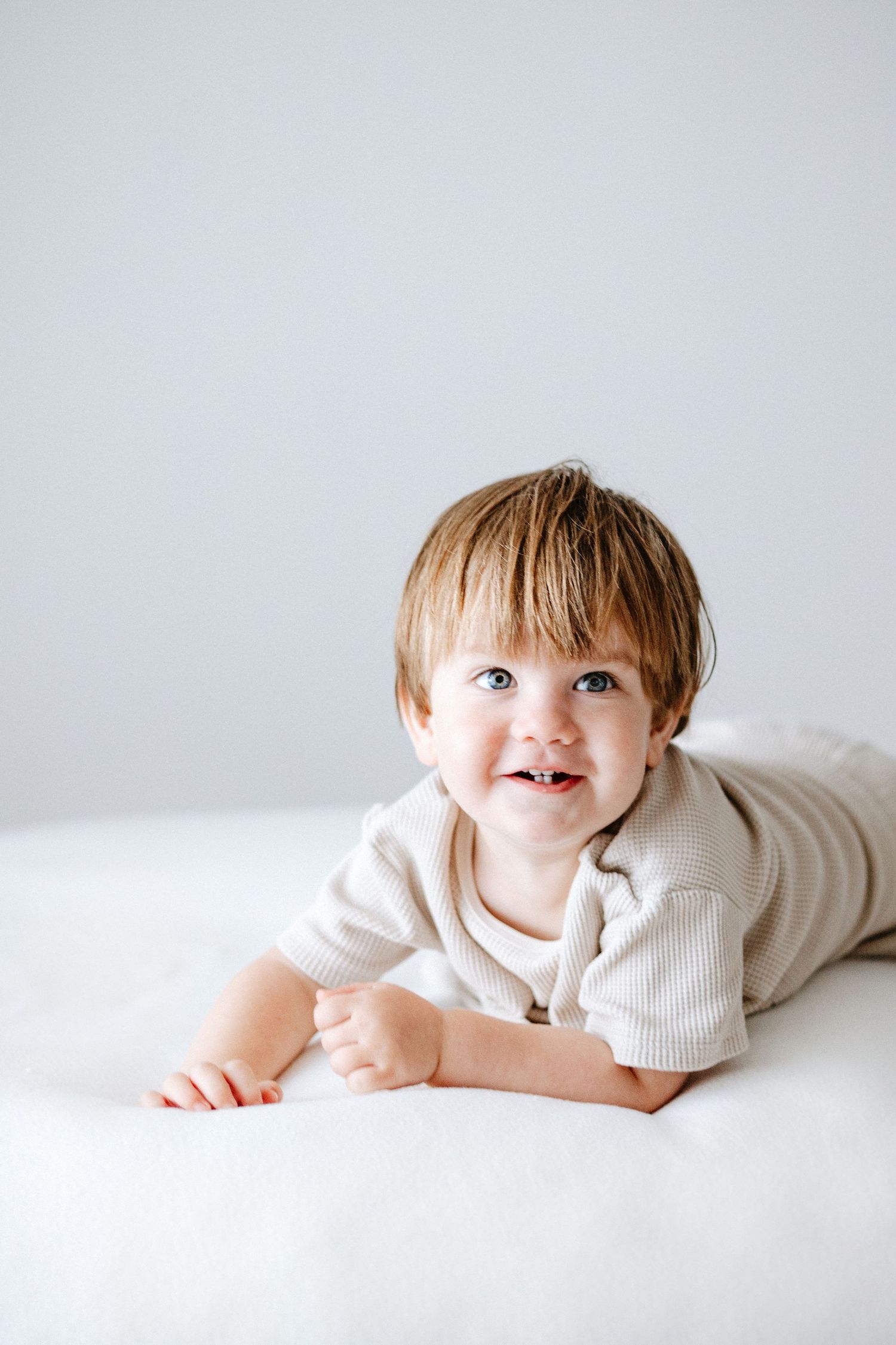 A young child in a white knit outfit lies on their stomach on a white surface against a light gray background.