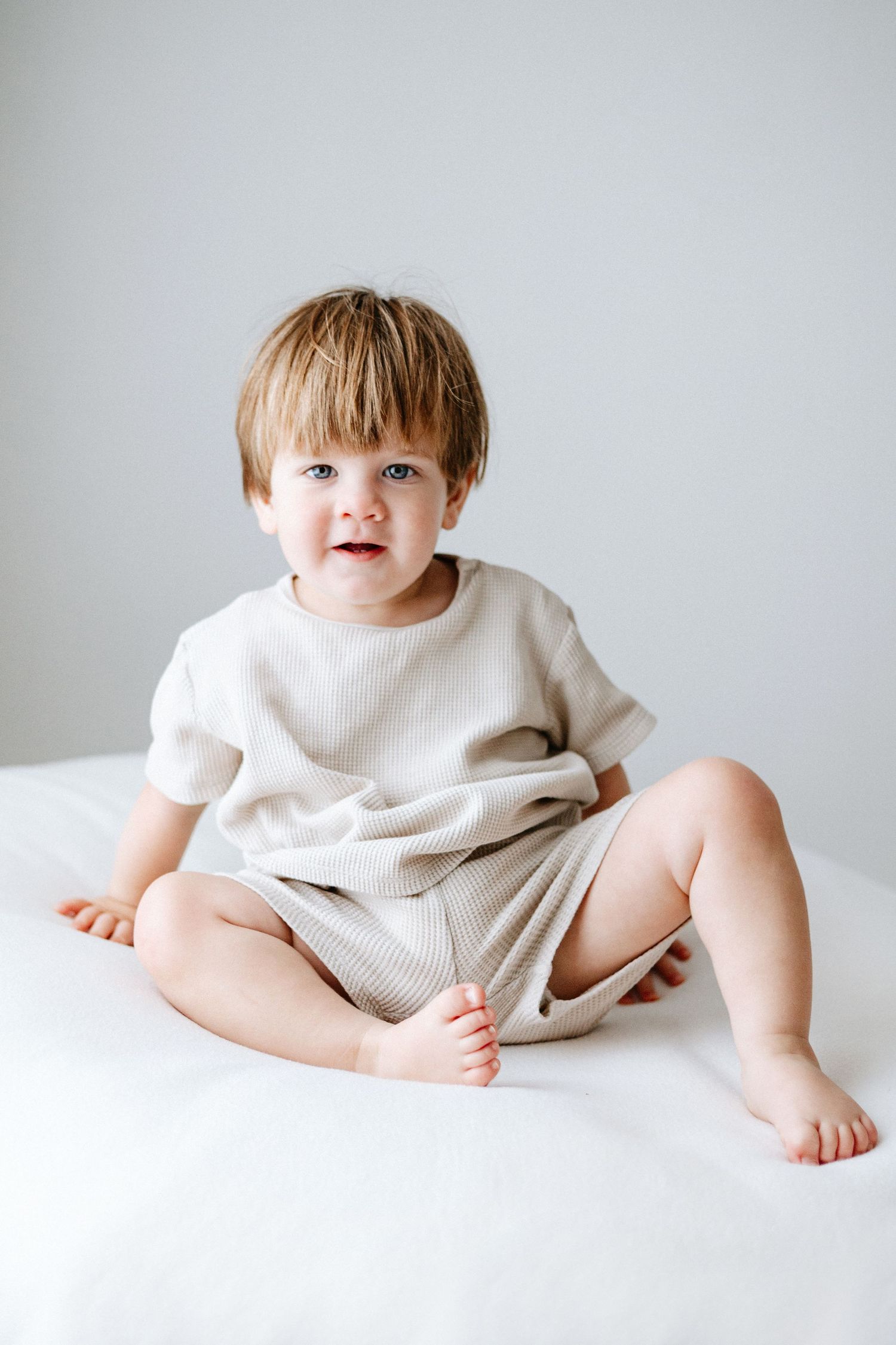Small child wearing white clothing sits on white backdrop in minimalist portrait series.
