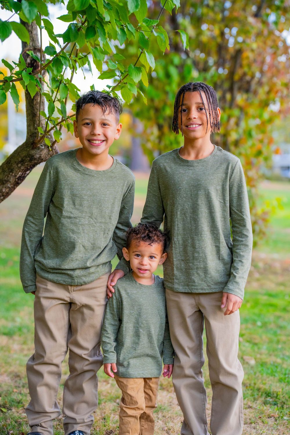 Family wearing matching olive green sweaters and khaki pants poses together outdoors under autumn trees.