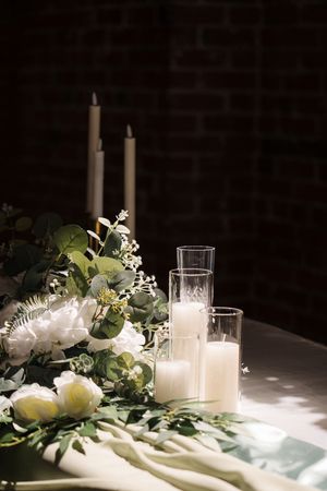 White pillar candles and delicate floral arrangements with greenery on a light-colored tablecloth.