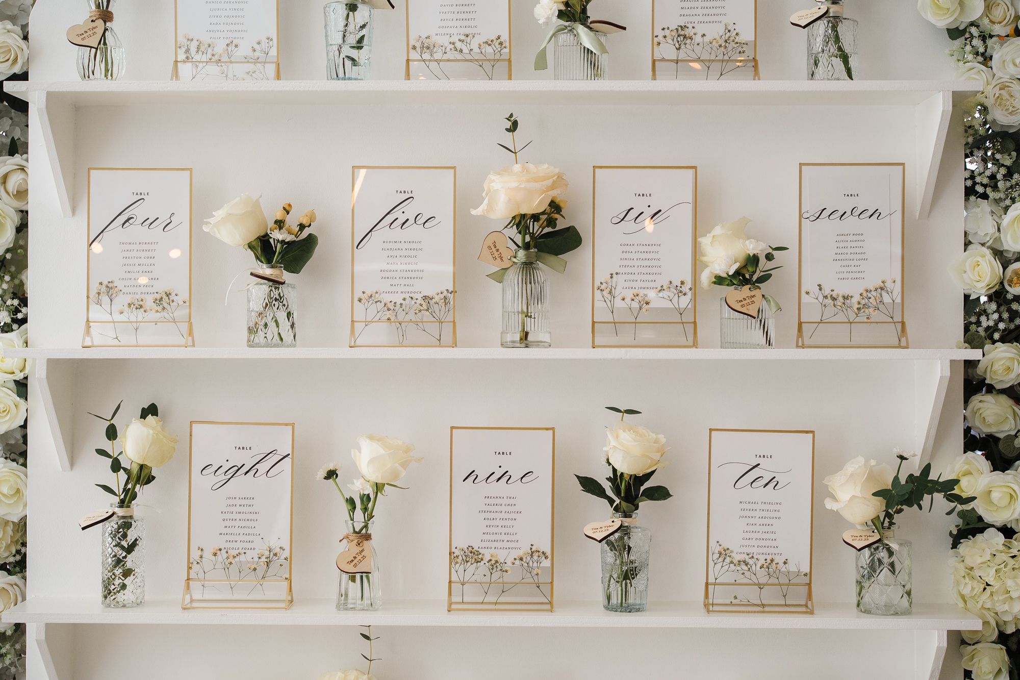 White floral table seating cards displayed on a wall with elegant calligraphy and rose decorations.