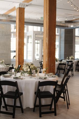 An elegant wedding reception venue with white round tables, black folding chairs and wooden support beams in an open space.
