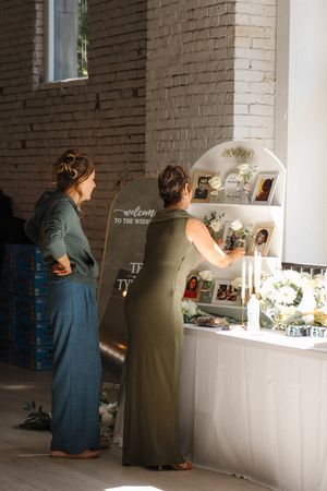 Two guests in long dresses view photos and decorations displayed at an elegant event setup against a brick wall interior.