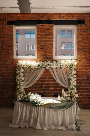 Elegant wedding reception backdrop with dreamy sheer gray drapes and white floral arrangements against exposed brick wall.