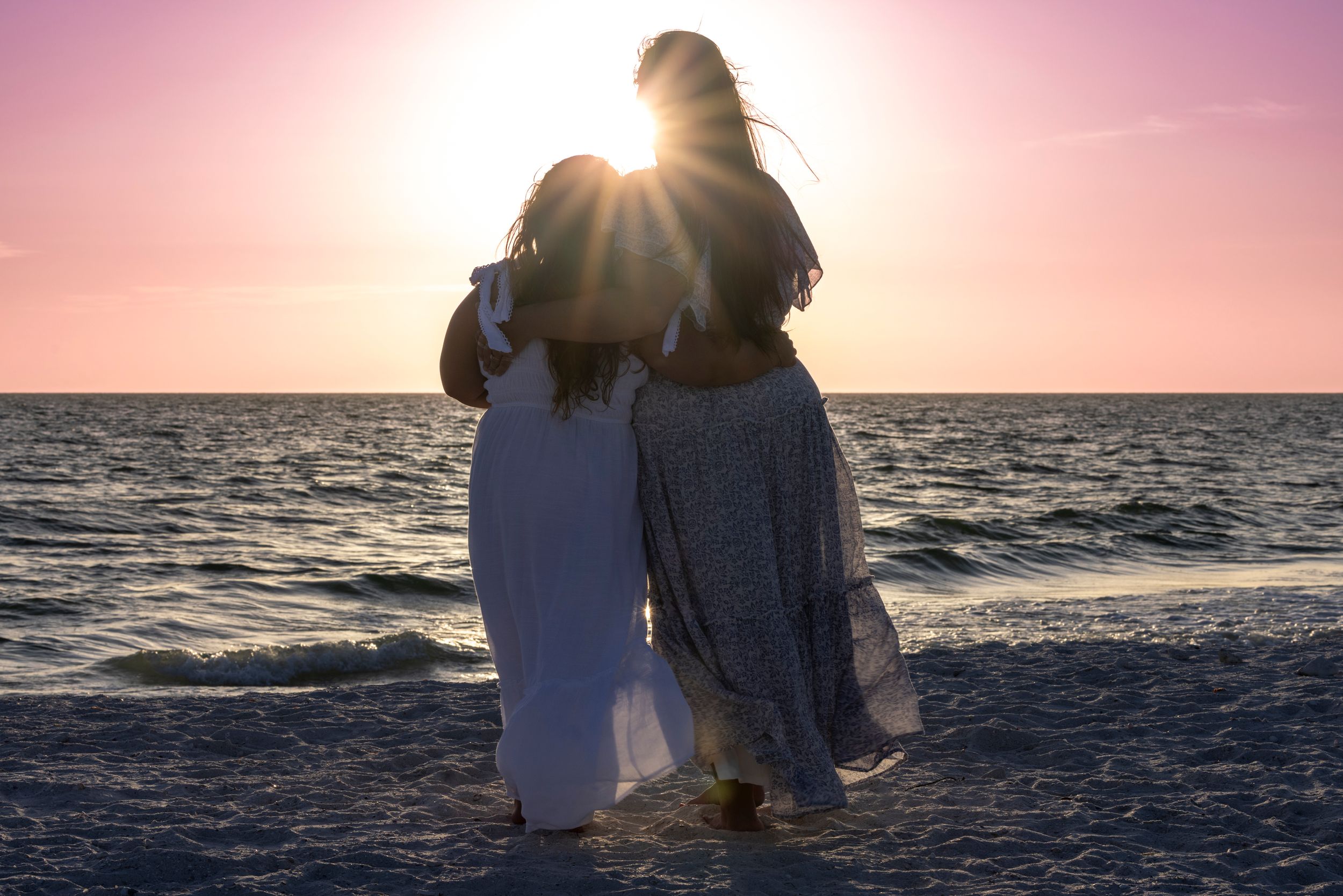 Two silhouetted people embrace while standing on a beach at sunset with sunlight creating a warm glow over the ocean.