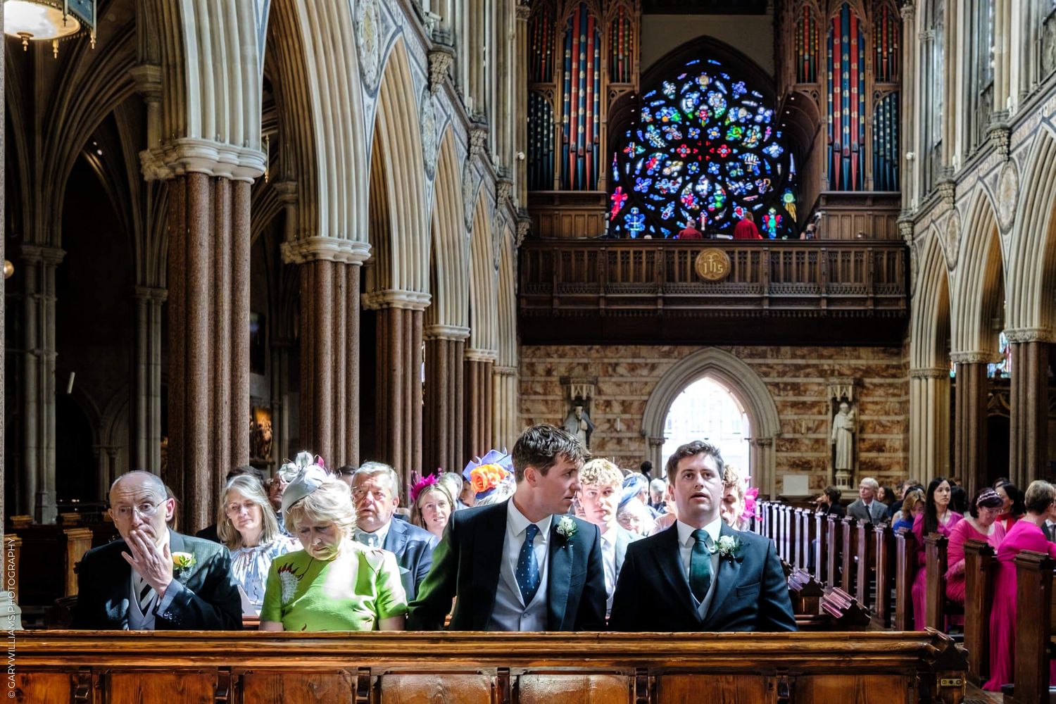 Wedding guests seated in wooden pews beneath stained glass windows and gothic arches of a historic church interior.