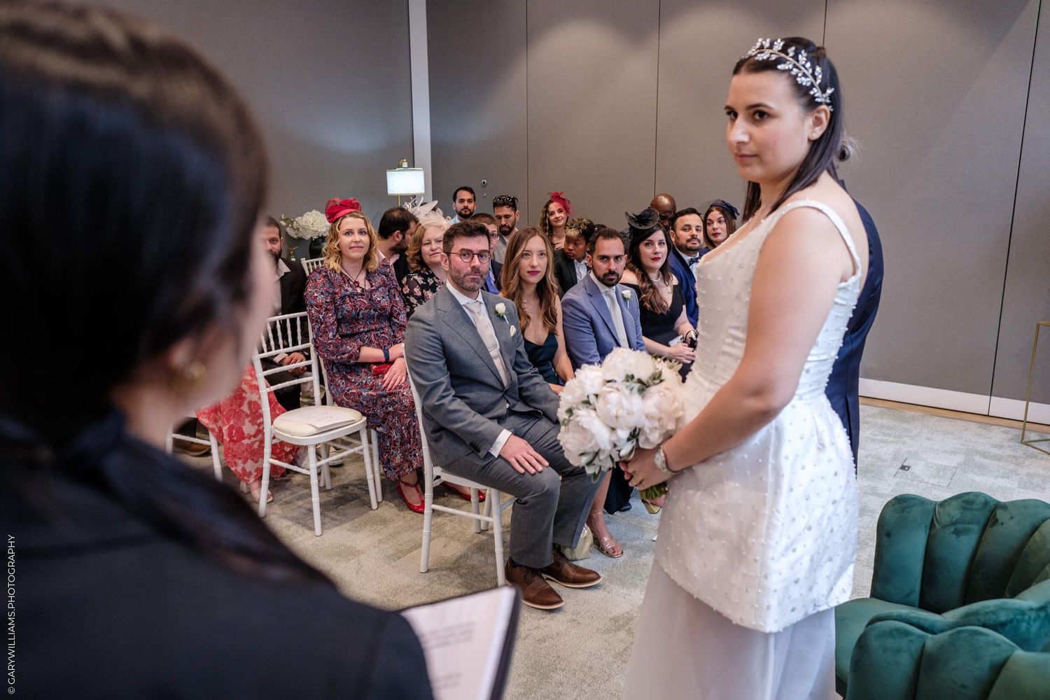 A bride in a white dress stands before guests during an indoor ceremony while an officiant conducts the service.