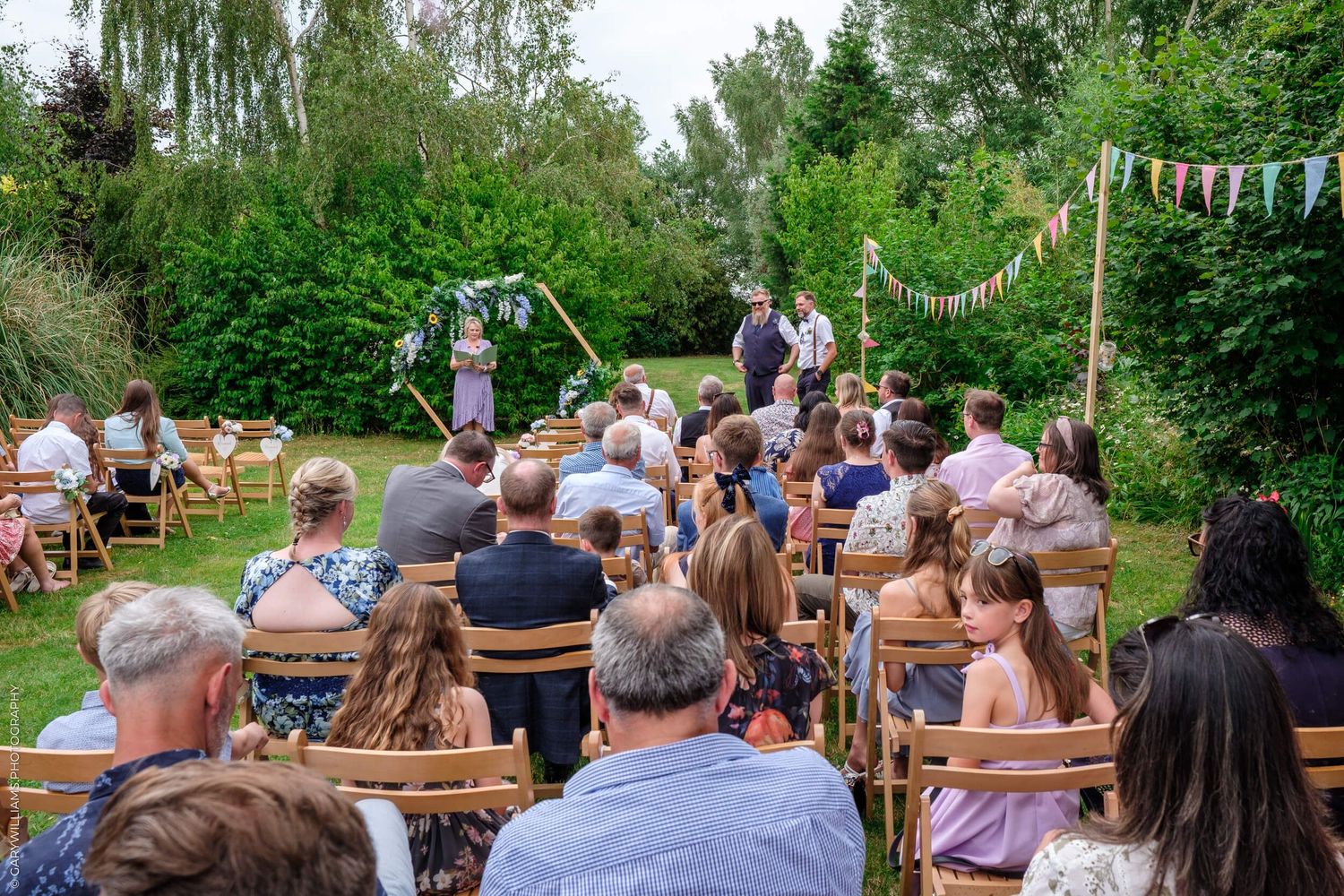 Outdoor garden wedding ceremony with bunting decorations and wooden chairs set up on the lawn.