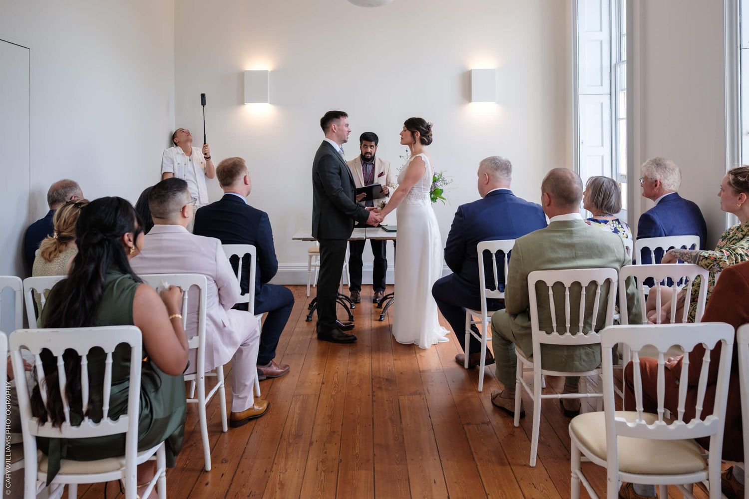 Wedding guests seated at tables in a bright, minimalist white reception venue with elegant chiavari chairs and pendant lights.